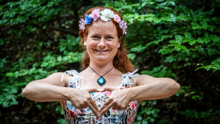 Marie-Gabriele Massa im Wald, formt mit ihren Händen ein Herz, trägt eine bunte Blütenkrone und ein Amulett – Ausstrahlung von Freude, Ruhe und Liebe