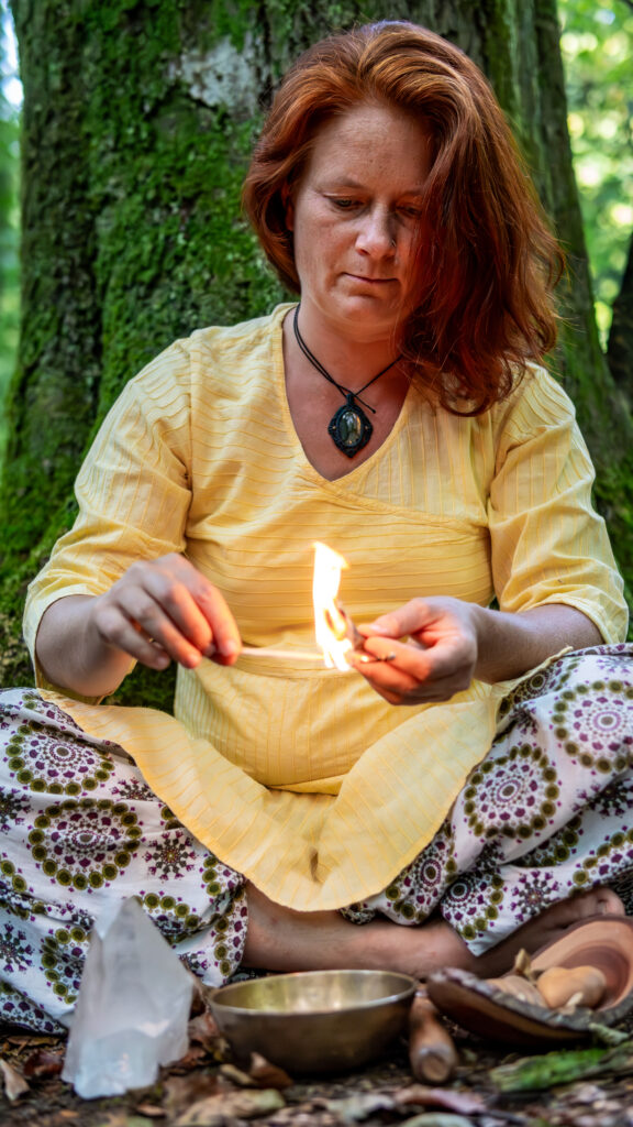 Marie-Gabriele Massa im Wald, sitzt im Schneidersitz vor einem riesigen Baum, zündet etwas an, mit Metallschale, Kristall und pflanzlichen Elementen – Naturverbundenheit und Ruhe