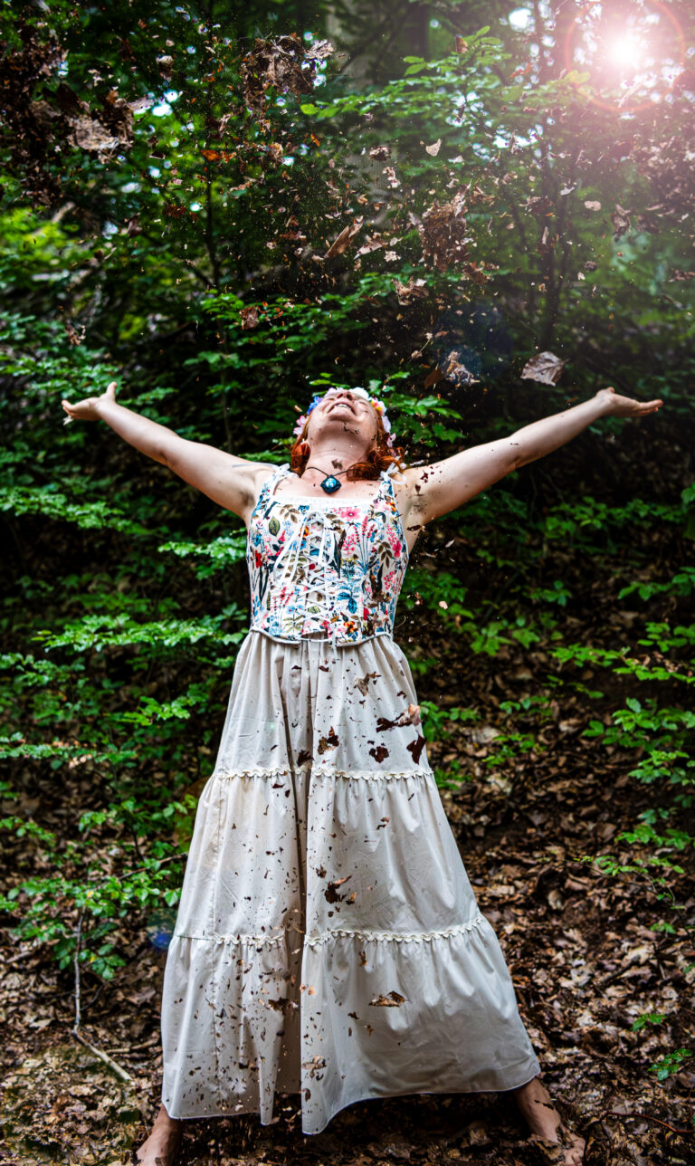 Marie-Gabriele Massa wirft Laub in die Luft – Ausdruck von Leichtigkeit, Freude und Naturverbundenheit, buntes Top, Kleid und Amulett im Wald