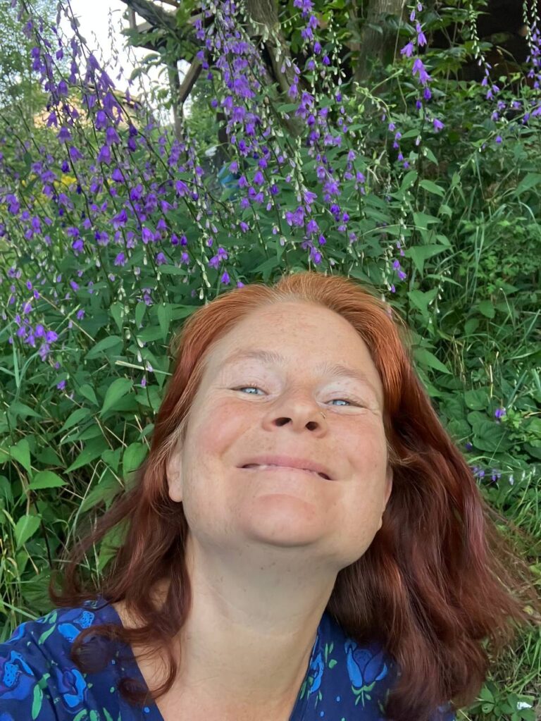 Marie-Gabriele Massa lacht fröhlich in die Kamera, im Hintergrund blühen Wildkräuter und lila Acker-Glockenblumen (Campanula rapunculoides)