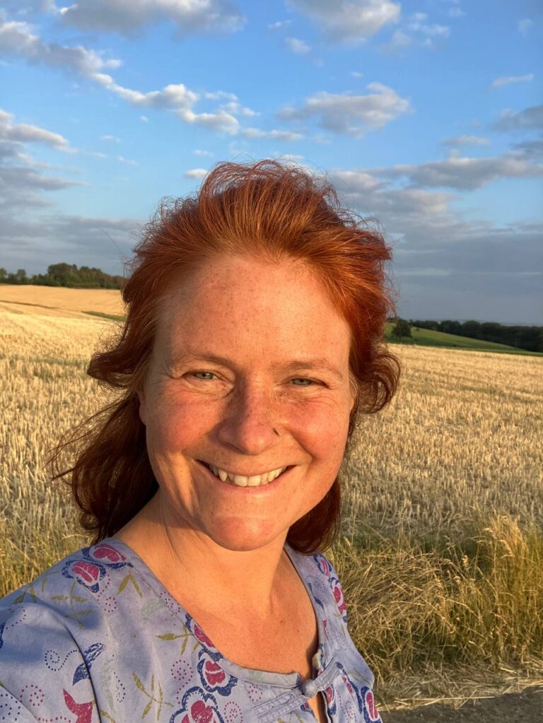 Marie-Gabriele Massa strahlend auf einem Feld bei Sonnenuntergang, mit Wolken, abgemähtem Feld und Wald im Hintergrund