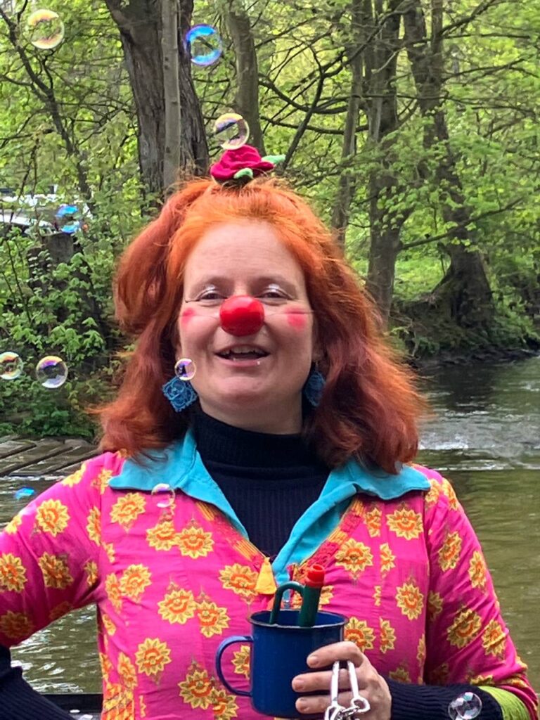 Marie-Gabriele Massa als Clown Tilotamma mit Seifenblasen, rotem Haar, lustiger Clownsnase, blauer Tasse und rosa Hemd – Naturcoaching und Clownerie im Saarland