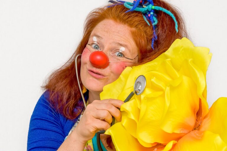 Marie-Gabriele Massa in Clownsverkleidung hält ein Stethoskop an eine riesige gelbe Blume – Clownerie in Merzig, Saarland